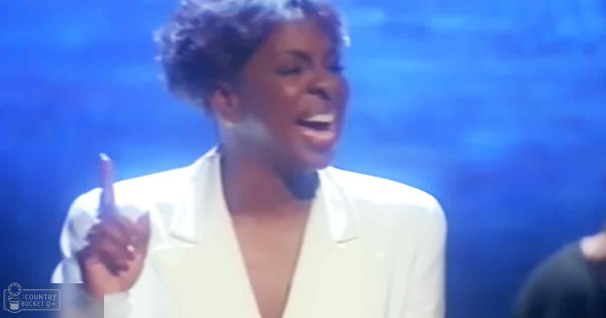 An adult female performer in a white blazer with 'Madly Odd' text, standing against a blue-lit backdrop likely indicating a stage or performance setting.