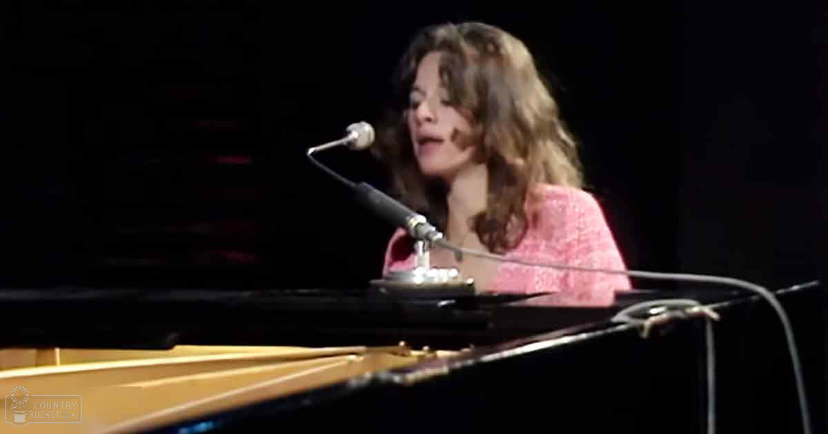 A young adult female in a pink patterned V-neckline with long sleeves, performing at an indoor music venue with stage lighting and a grand piano.