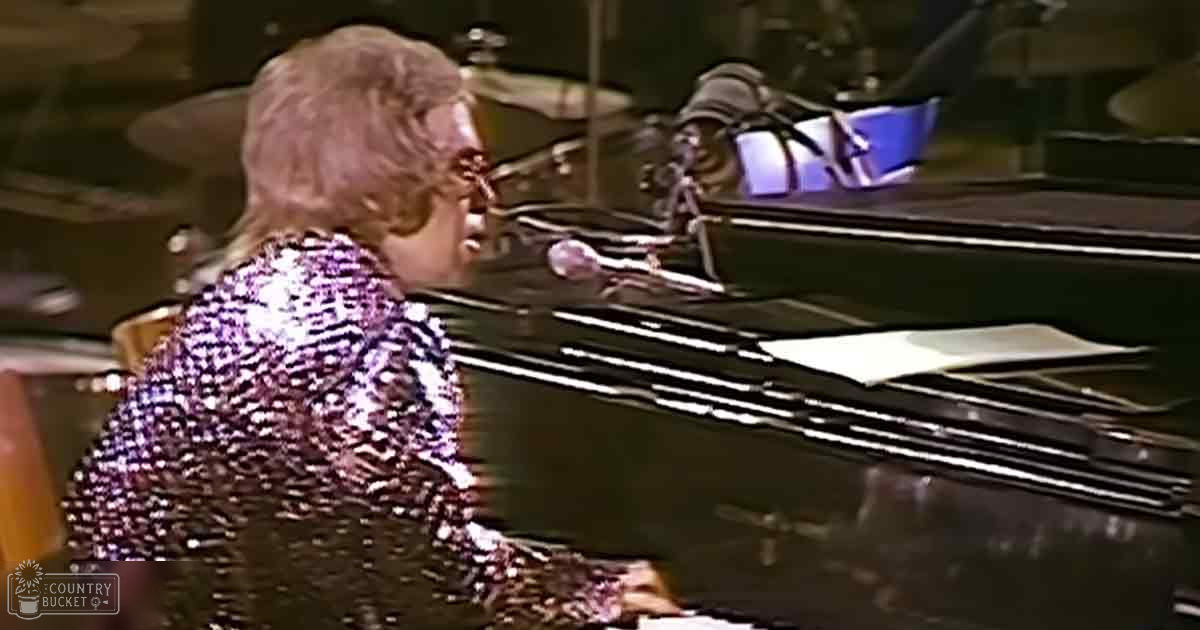 A middle-aged male singer in a dark 1970s-80s style, shiny sequined jacket with reflective material, performing at an indoor concert venue with stage lighting and a grand piano.