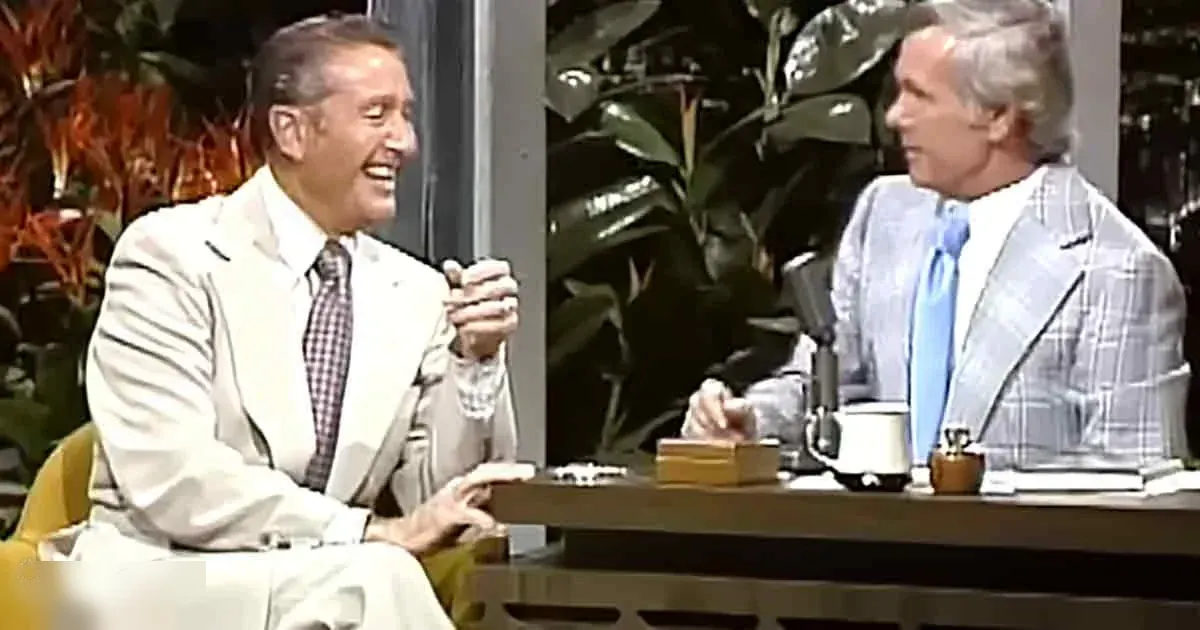 Talk show setting with two adult males engaged in conversation, relaxed attire, indoor studio with green plants and past decade ambiance.