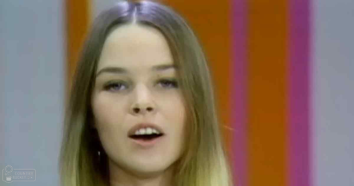 A young adult female wearing a light-colored, solid top with modern casual style indoors against an orange and pink striped background.