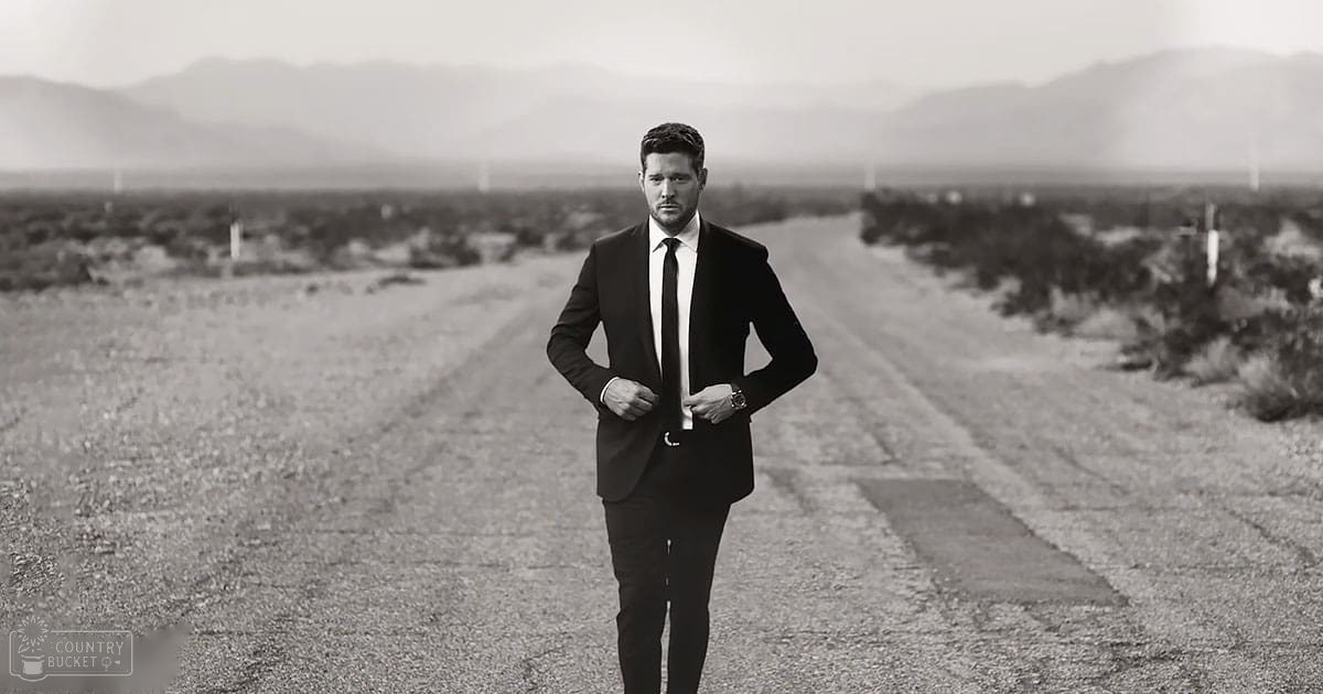 Adult male in formal black suit and white shirt on open desert road under hazy sky, suggesting late afternoon or early evening conditions.