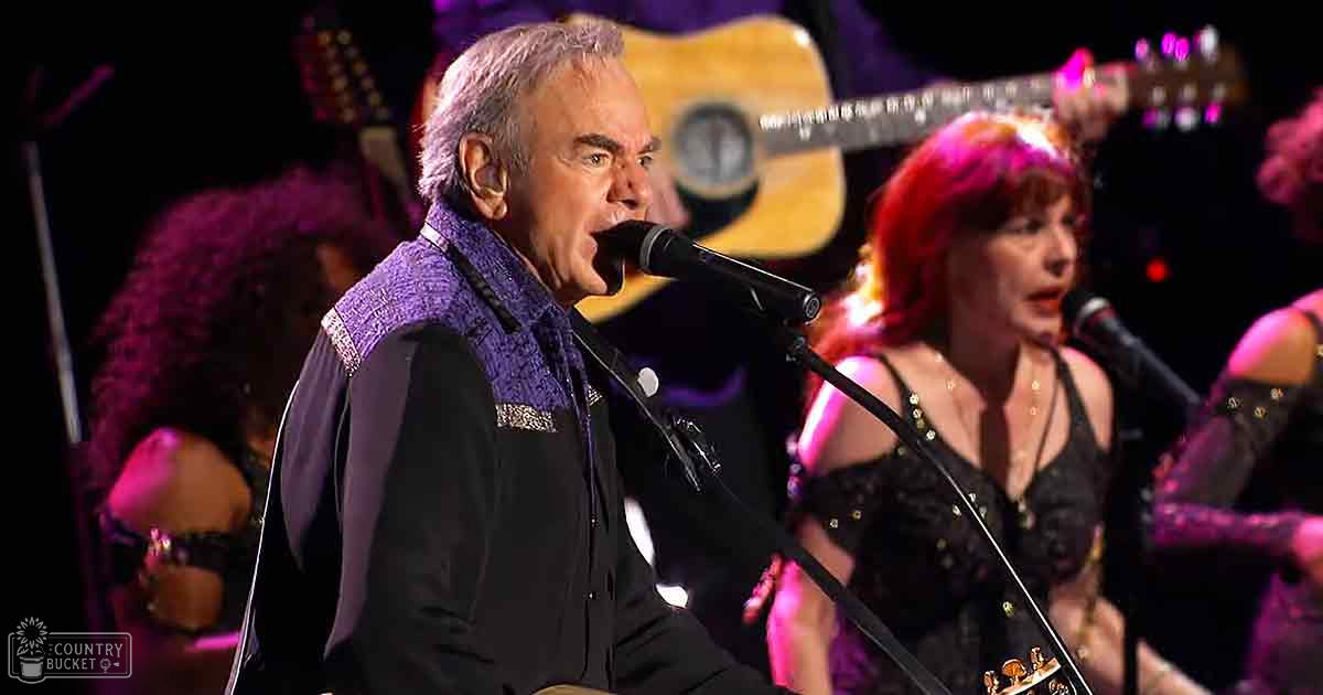 An older adult male singer performing on a concert stage with warm stage lighting and other band members in the background.