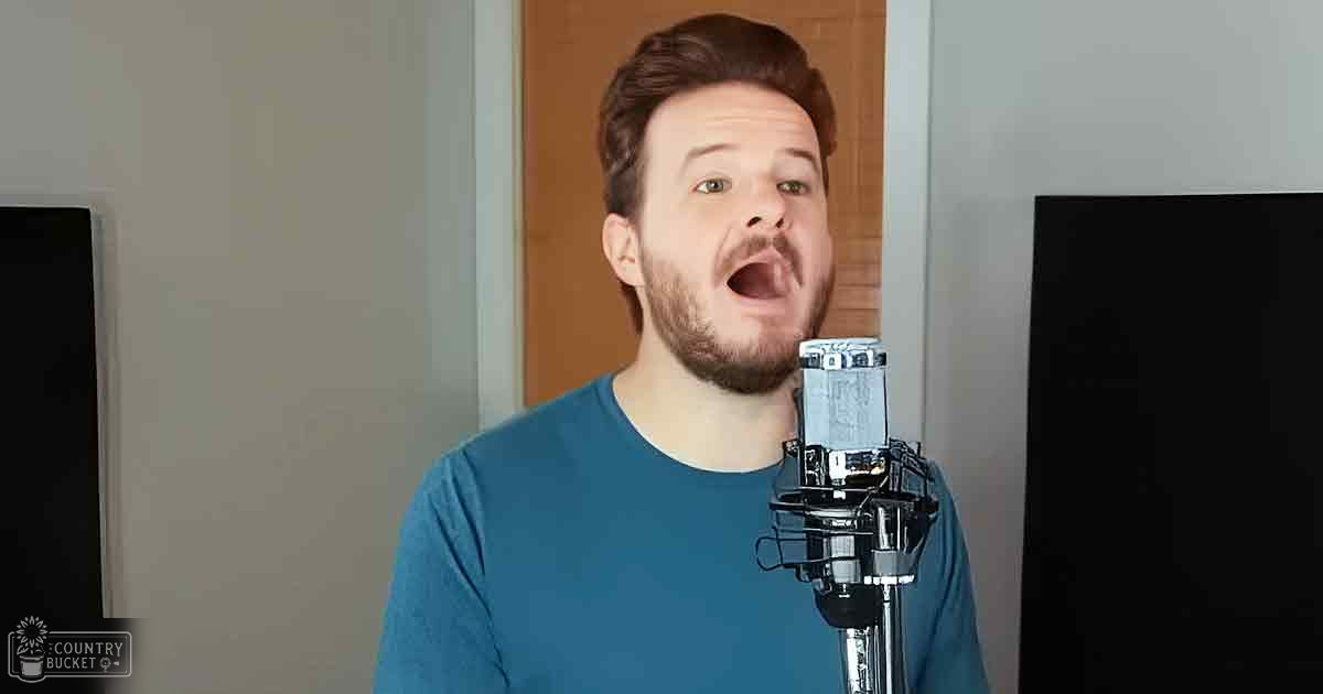 A young adult male wearing a blue, relaxed fit long-sleeved shirt indoors with a microphone and door in the background.