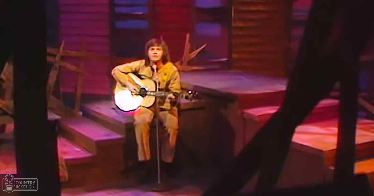 A young adult male with long hair plays guitar on wooden steps, suggesting a live performance setting.