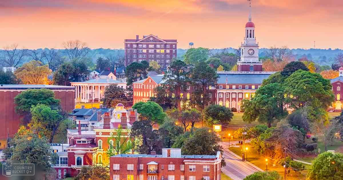 A historic university campus at dusk with red-brick buildings, white columns, and a clock tower surrounded by trees and hills in an urban environment with warm sky hues indicating sunrise or sunset.
