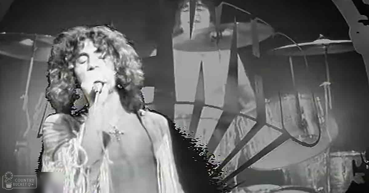 A young adult male singer wearing a long-sleeved fringe garment with a cross pendant, singing into a microphone on an indoor stage.