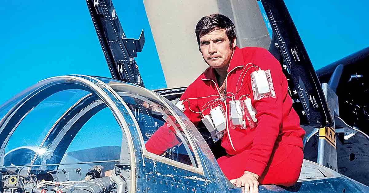 An adult male pilot in a red flight suit with white patches, wearing wristwatch, inside an aircraft cockpit under blue sky and part of an aircraft hangar.