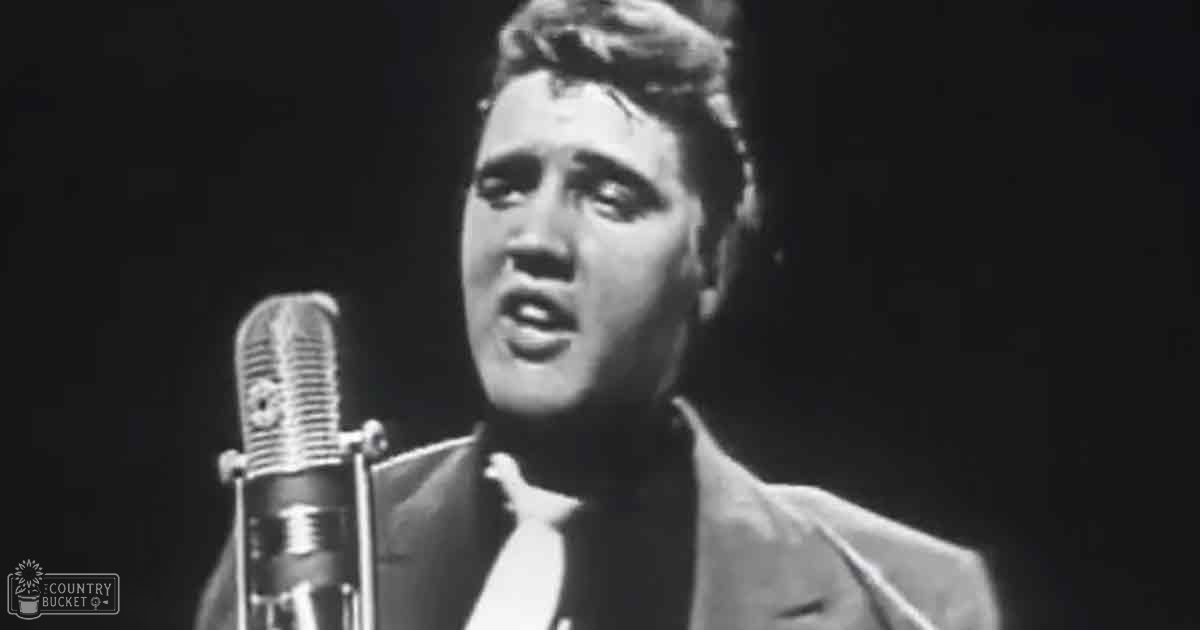 A young adult male performer in a well-fitted suit jacket, white shirt, and dark tie stands before a microphone at what appears to be a concert or recording session. The vintage-style microphone has 'Madly Odd' printed on it.