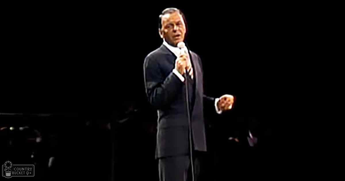 A middle-aged male performer wearing a well-fitted dark suit and white shirt, holding a microphone in an indoor concert setting with dim stage lighting.