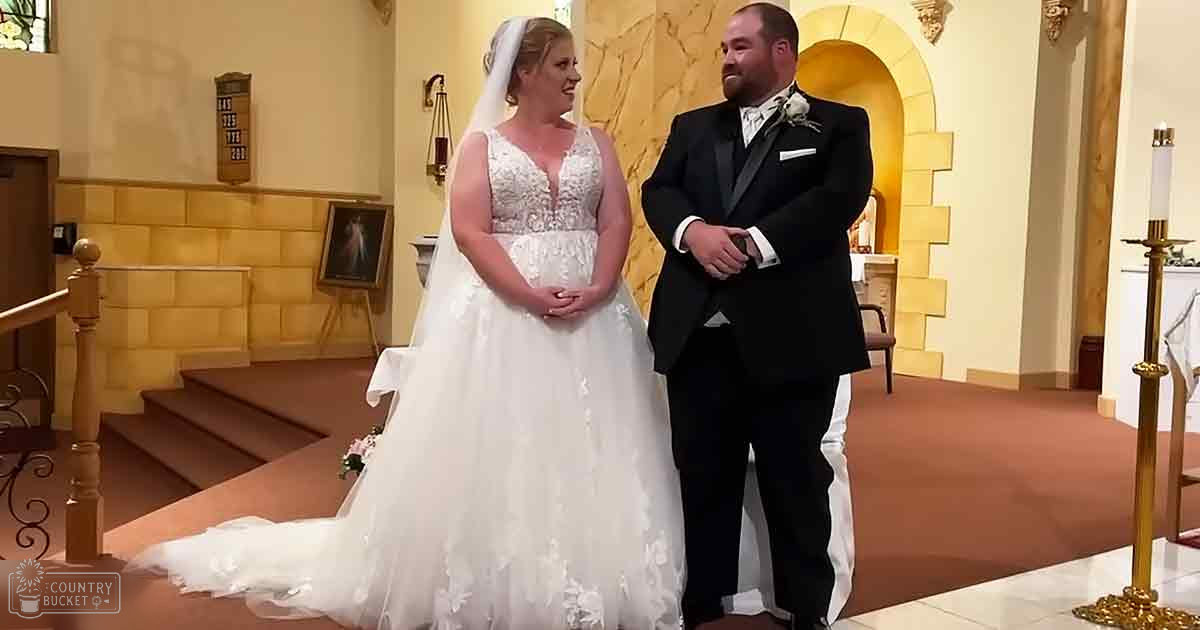 A bride and groom in a church, surrounded by ornate religious decor; the bride wears an intricate white wedding dress with floral lace detailing, while the groom dons a black tuxedo with a boutonniere.