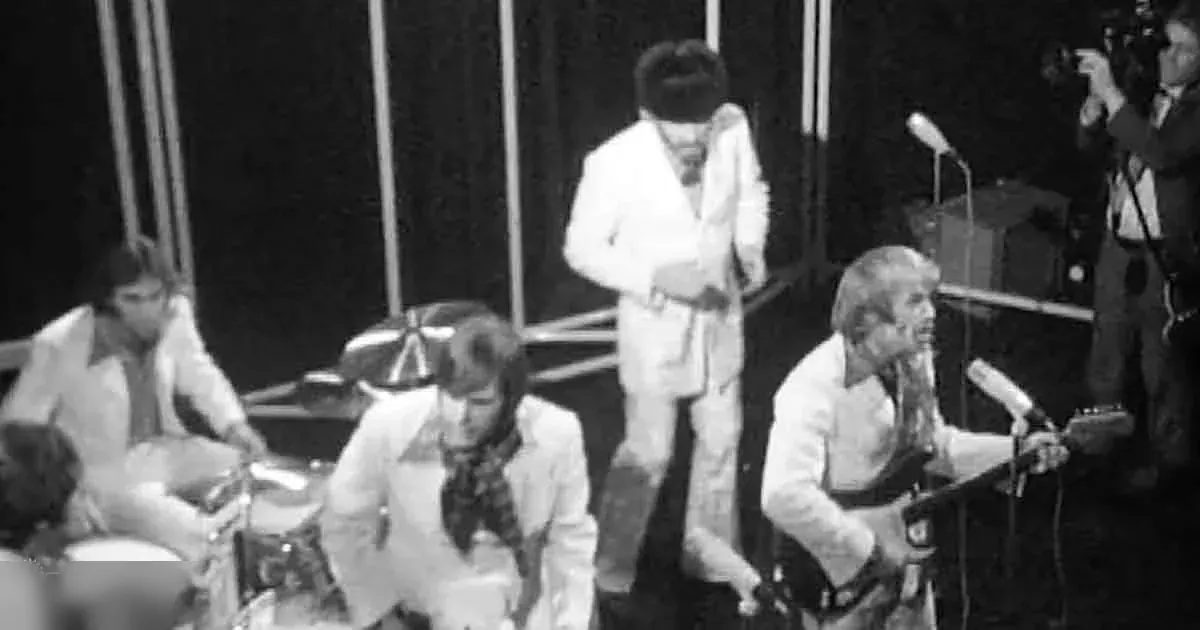 A musical performance from the 1960s-1970s era with a young adult male drummer, another young adult male singer with microphone, and a middle-aged man playing keyboard on stage. The setting is an indoor concert venue or studio with curtains in the background.