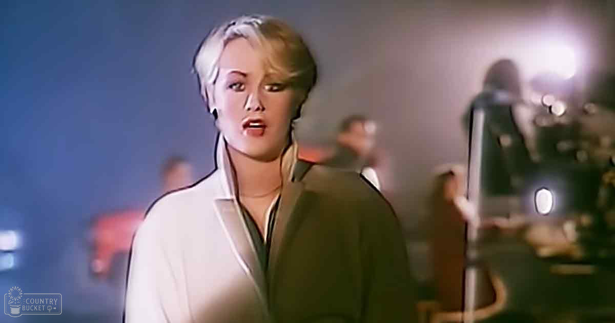 A young adult female wearing a white blazer with black trim, green shirt underneath, and possibly a necklace or choker, standing indoors under artificial dim lighting.