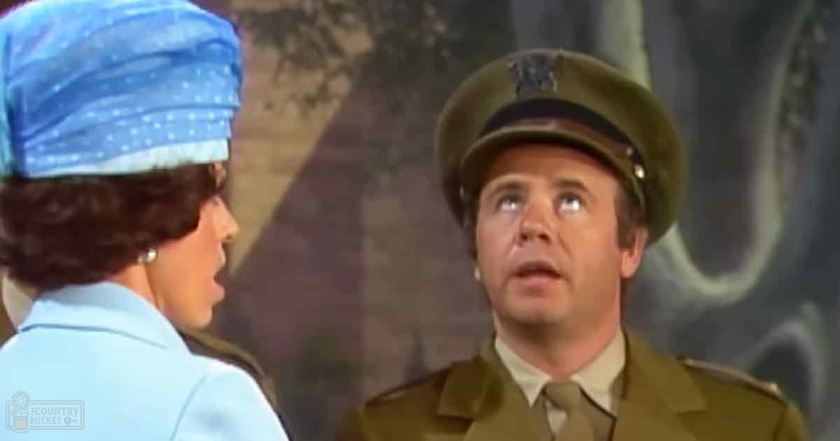 A man wearing a mid-20th century military uniform and pearl earrings stands indoors with an adult female wearing a light blue blouse and polka dot headscarf, in muted tones under diffused lighting.