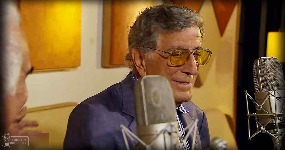 Older adult male in a recording studio, wearing a dark blue suit with a white shirt and patterned tie, surrounded by microphones and soundproofing materials.