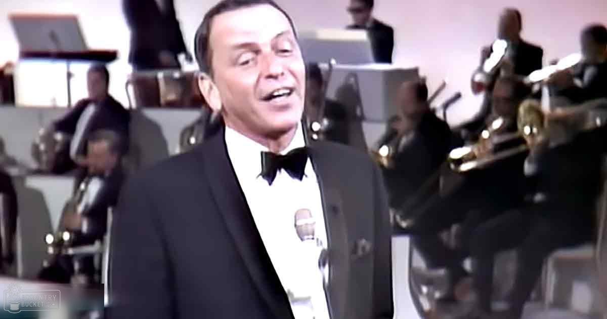 A middle-aged male adult wearing a black tuxedo with a white shirt and bow tie, standing on what appears to be a stage in an indoor venue likely used for performances.