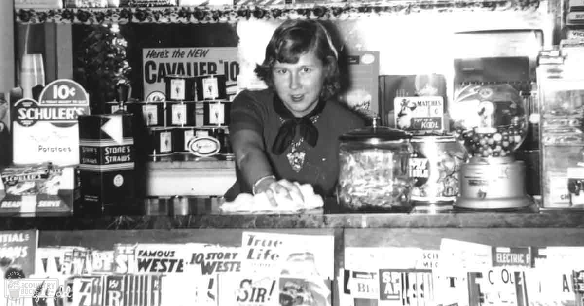 A young adult female wearing a dark-colored top with long sleeves and a bow at the neckline, standing in a mid-20th century store setting filled with vintage products.