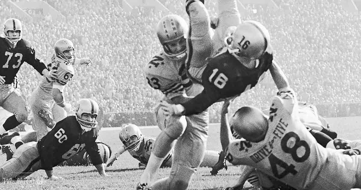 A football game in progress at a stadium, with players in black and white jerseys with numbers visible, surrounded by spectators under bright daylight.