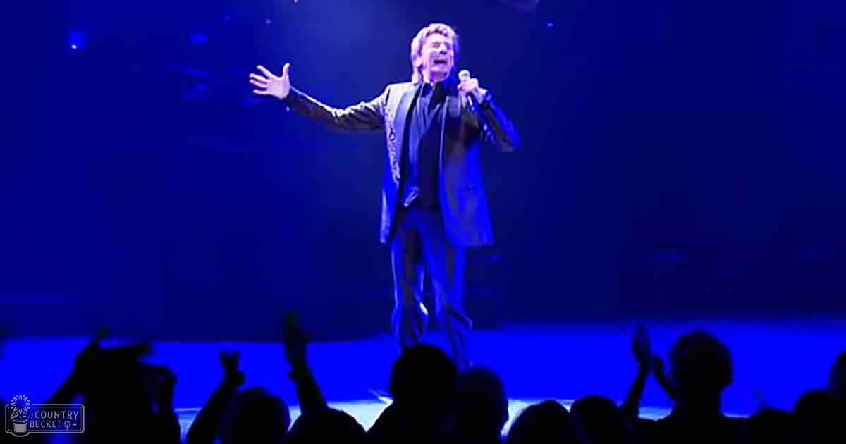 An adult male singer in a dark suit jacket and pants holds a microphone on stage at a concert venue under blue lighting.