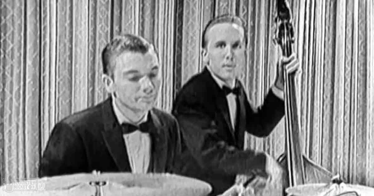 A young adult male singer in a dark suit jacket with a white shirt and black bow tie, performing on stage alongside an older adult male, both dressed similarly.