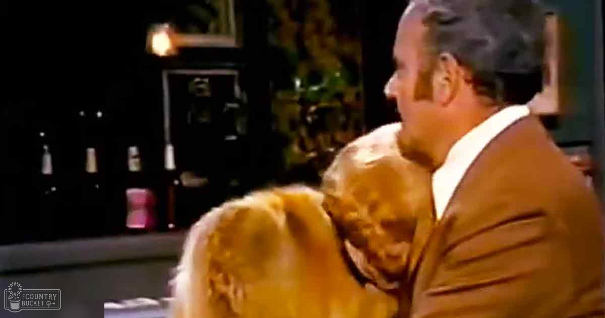 An older adult male holding a toddler girl in an indoor setting with a golden retriever dog nearby and personal decor visible.