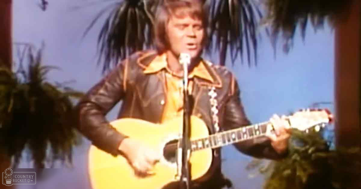A middle-aged person in a 1970s-style leather jacket plays a yellow acoustic guitar on an indoor stage with artificial palm leaves, suggesting a tropical or beach theme. The setting is bright and even-lit, typical of performance venues from that era.