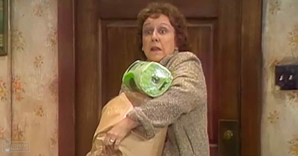 A middle-aged adult female wearing a grayish-brown sweater and beige pants, standing in an indoor room with wooden doors and floral wallpaper; the focus is on a green plastic bottle wrapped in cloth or paper.