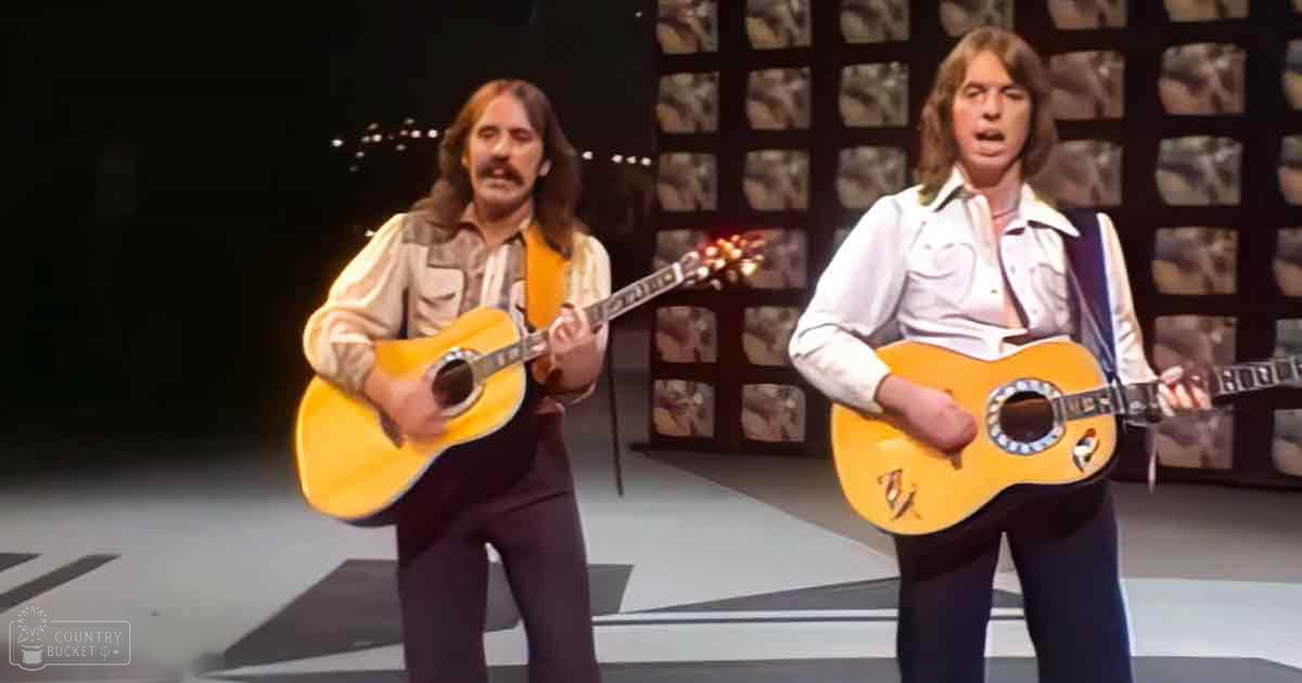 Two male performers in a musical performance scene, wearing patterned shirts and holding guitars; the setting is an indoor stage with square panelled walls.