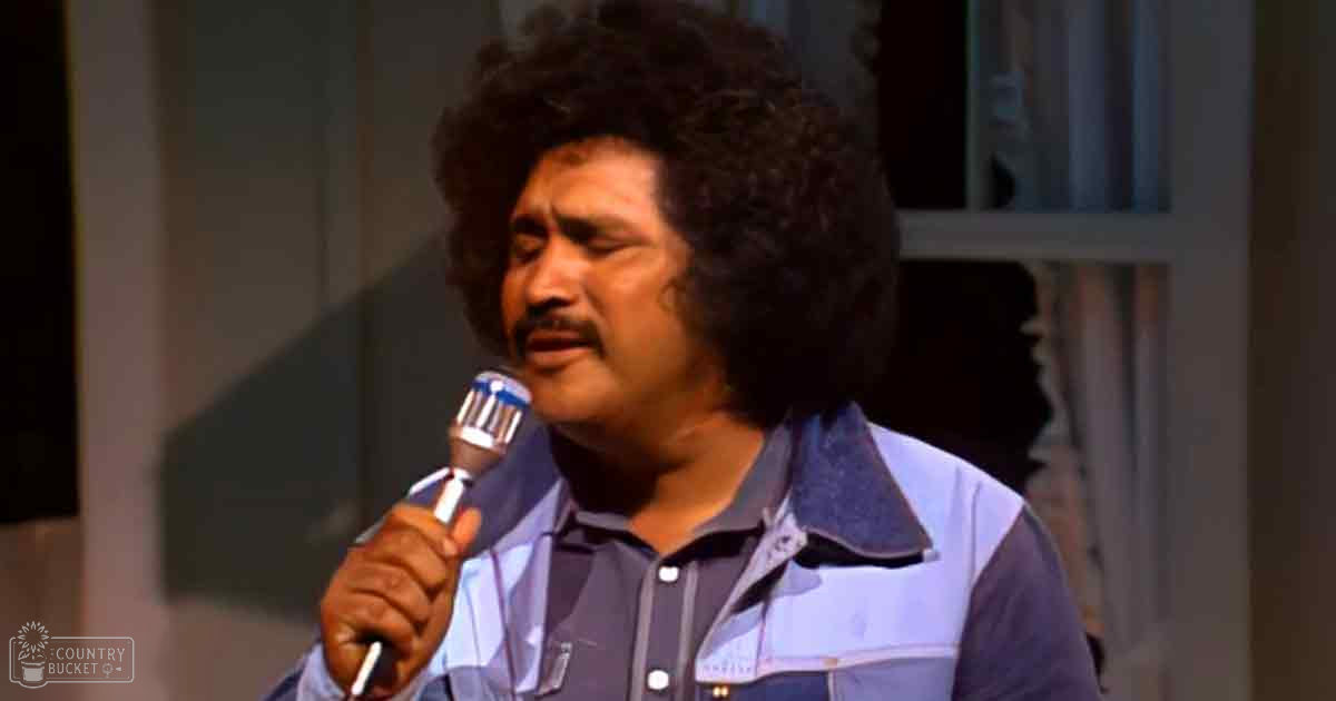 A middle-aged adult male in a relaxed denim jacket and light blue shirt indoors, holding a microphone with a vintage style, suggesting the era is likely from 1970s to early 1980s.