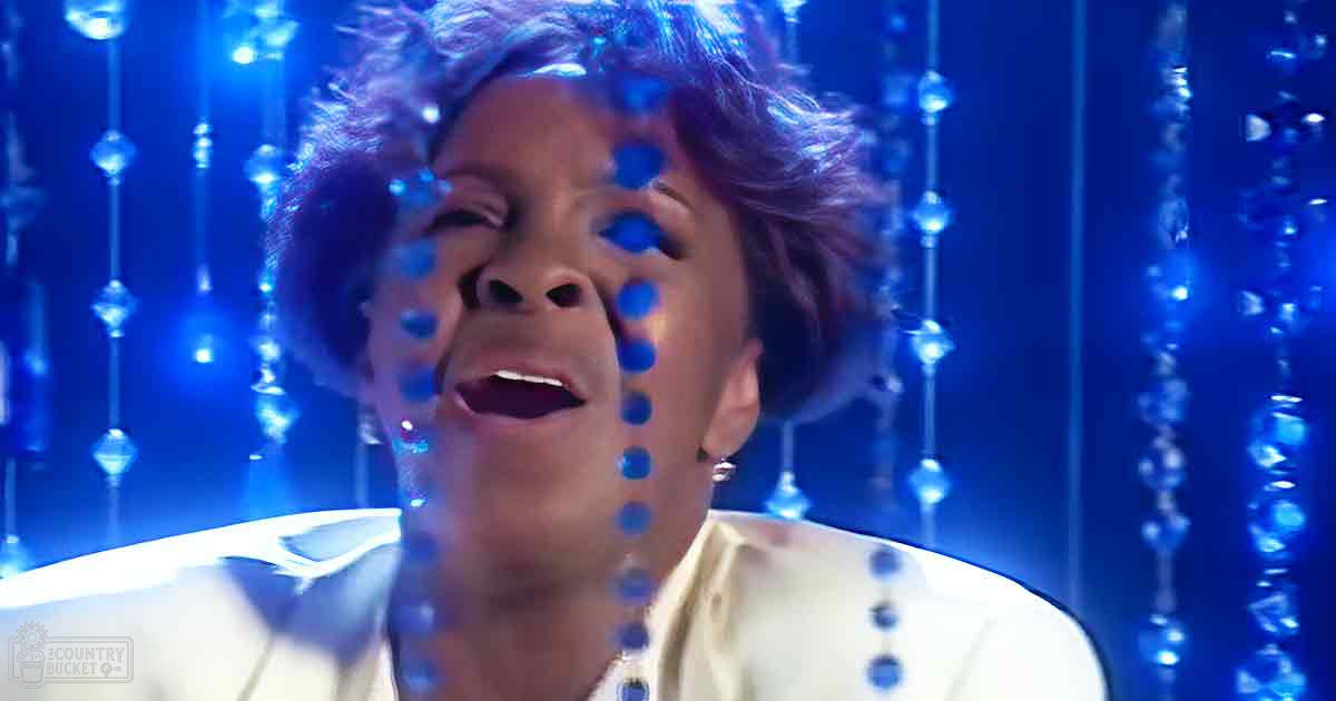 A middle-aged adult woman in a studio setting with reflective water droplets, wearing a white blouse and earrings under blue lighting.