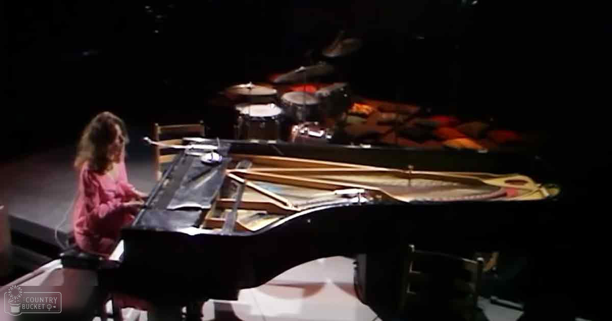A young adult female plays a grand piano in what appears to be a concert hall with sheet music stands and stage lighting typical for evening performances.