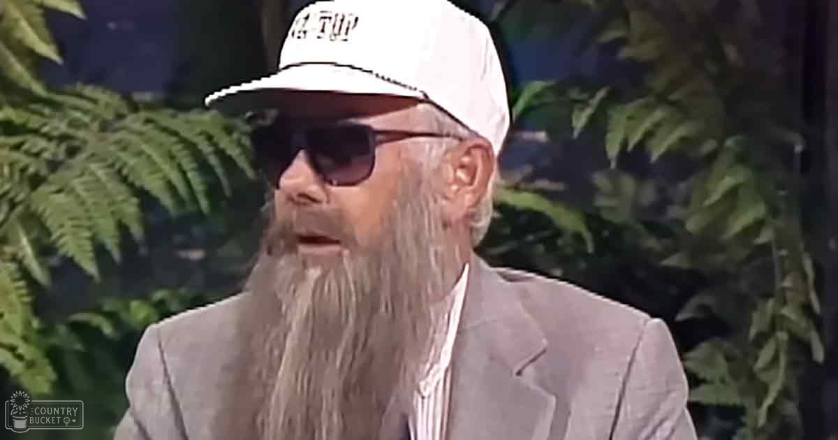 An older adult male wearing a light gray suit jacket with '4 TOP' on his white cap, dark sunglasses, in an outdoor garden setting.