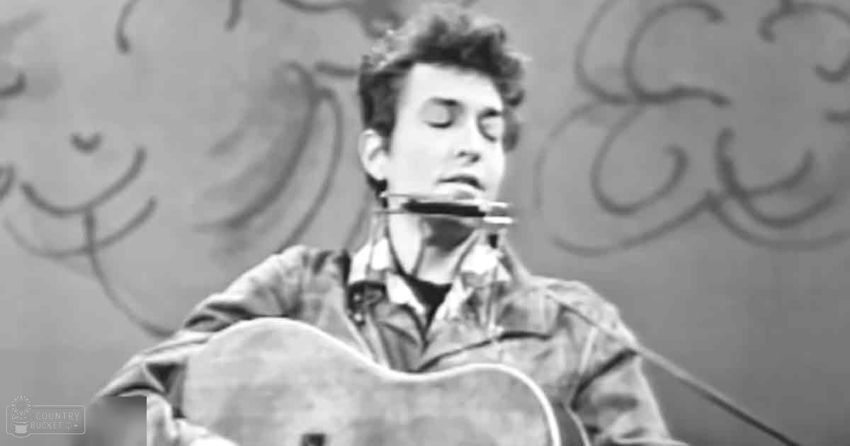 A young adult male playing a guitar and harmonica indoors with a graffiti-covered wall behind him.