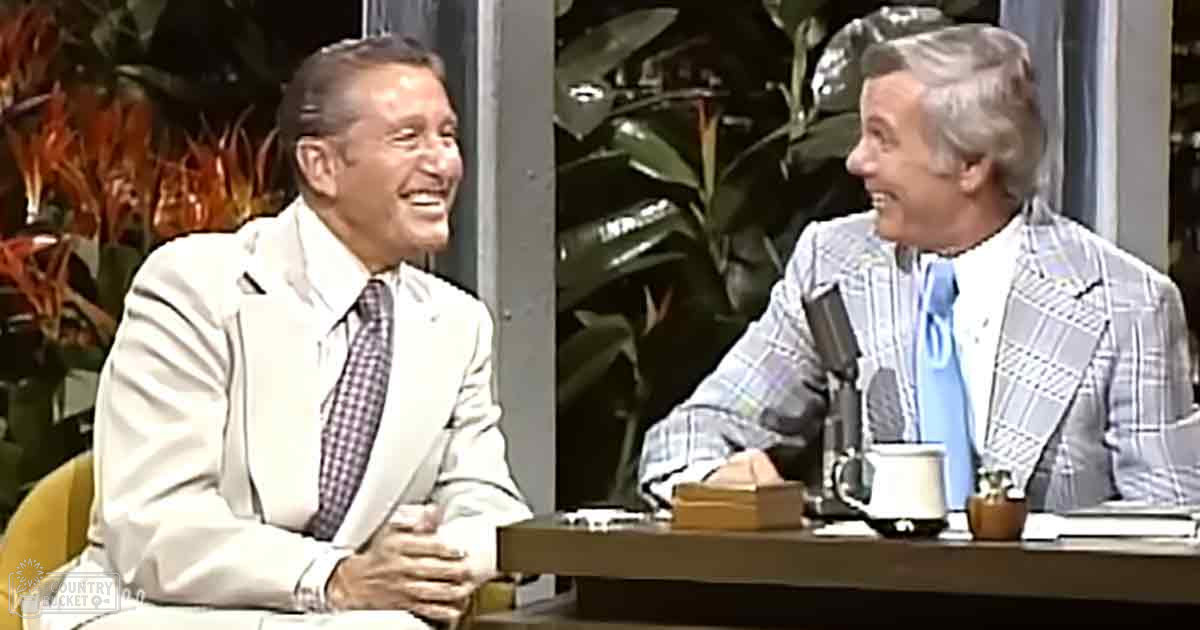 Two older adults in a talk show setting with indoor plants and even lighting.