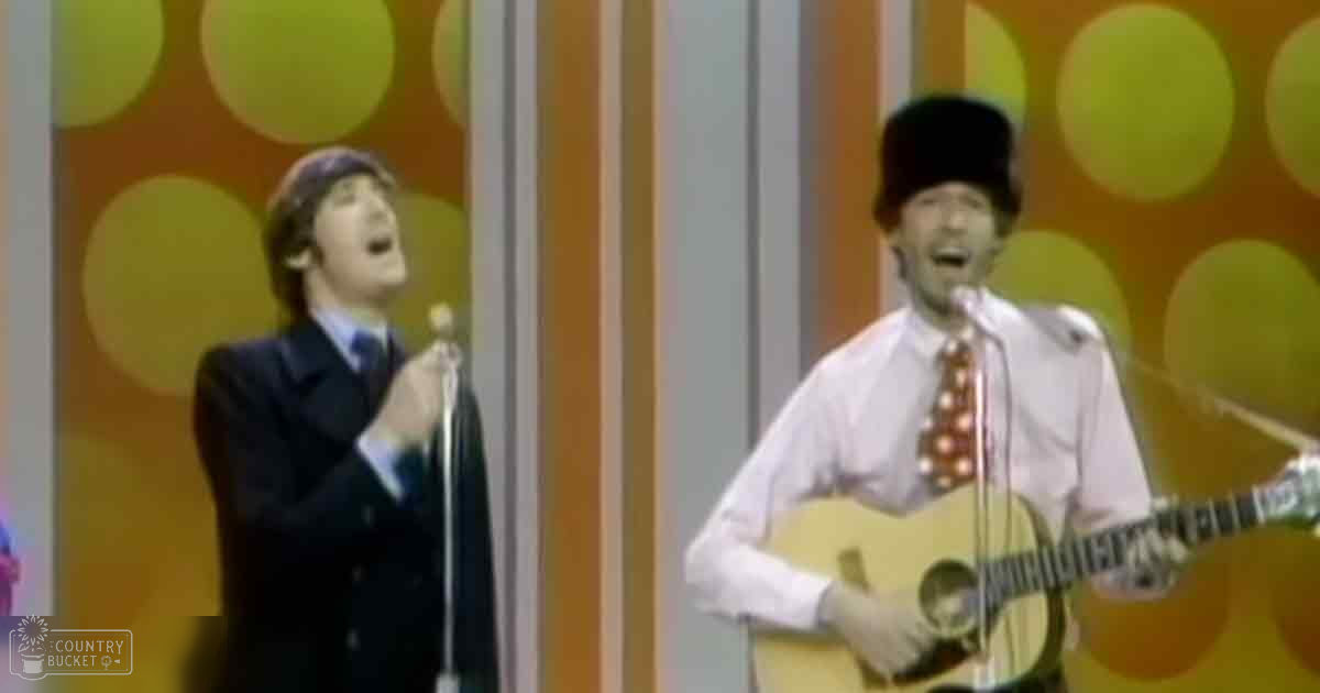 A mid-20th century musical performance scene with a young adult male singer holding a microphone stand next to another young adult male wearing a white shirt and patterned tie, both on stage against an orange-yellow-white striped backdrop.