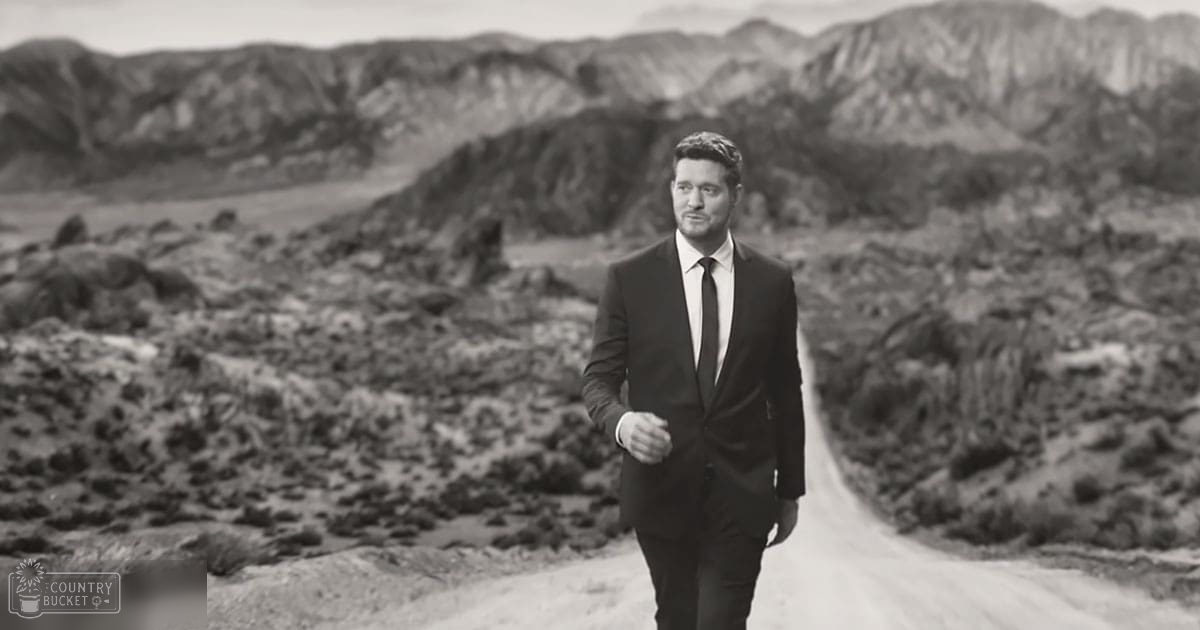 Adult male in a tailored dark suit jacket and trousers standing outdoors in a desert landscape with rocky terrain and mountains, early morning or late afternoon light.