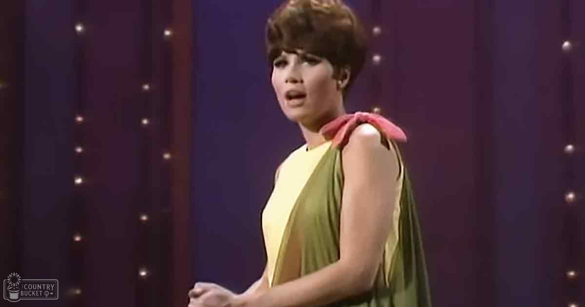 A young adult female in a flowy sleeveless dress with draped fabric, standing on an indoor stage or performance area.