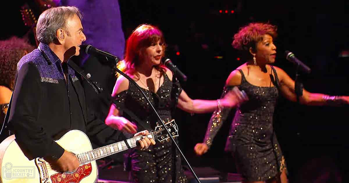 Three adult musicians in concert attire performing under stage lighting, with a white and red-gold patterned guitar as the focal point.