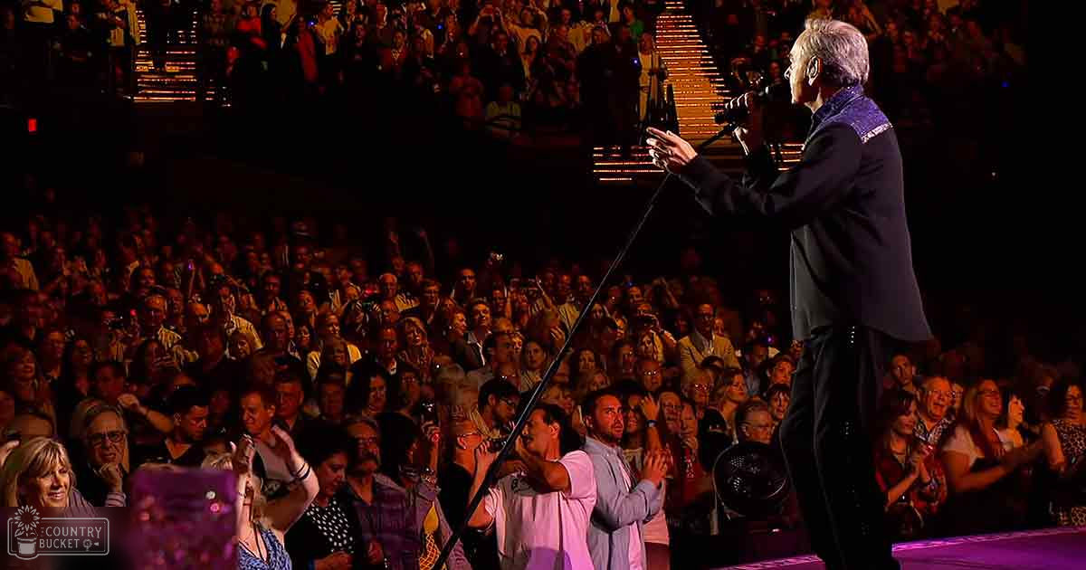 A concert scene indoors at an arena or theater; audience members hold up phones and cameras, dressed in casual attire with bright stage lighting illuminating performers.