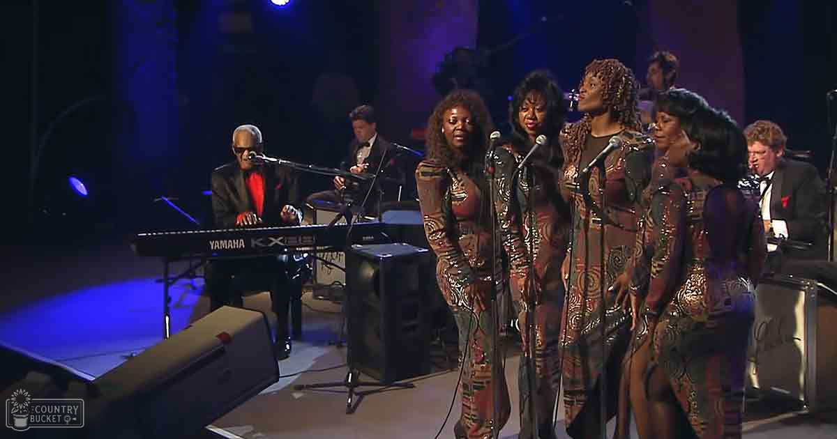 Adult male in dark suit plays keyboard while singing; four adult females wear matching patterned dresses with earthy tones, also sing and play microphones.