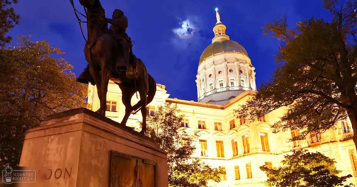 A nighttime view of a statue of a man on horseback at the Georgia State Capitol building, illuminated by warm yellows with deep blues and browns from trees and statue base in the foreground.