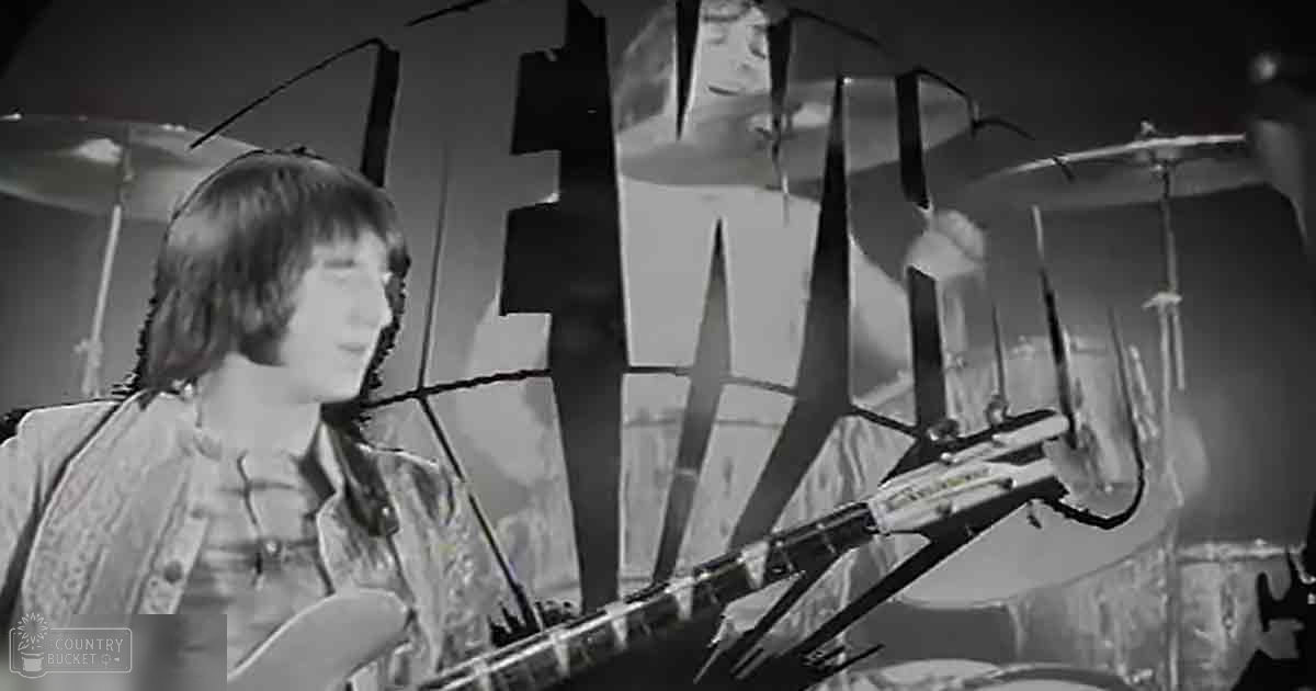 A young adult male playing guitar at a music venue with 'The Who' sign in the background, wearing a light-colored paisley shirt from the late 1960s or early 1970s.