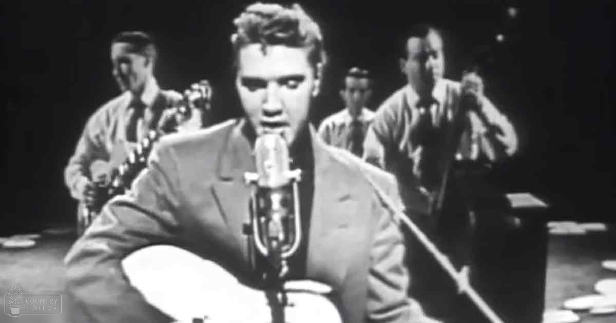A young adult male singer in a light-colored suit jacket and white pants stands before a microphone on stage with musicians playing guitars, all under dim indoor lighting.