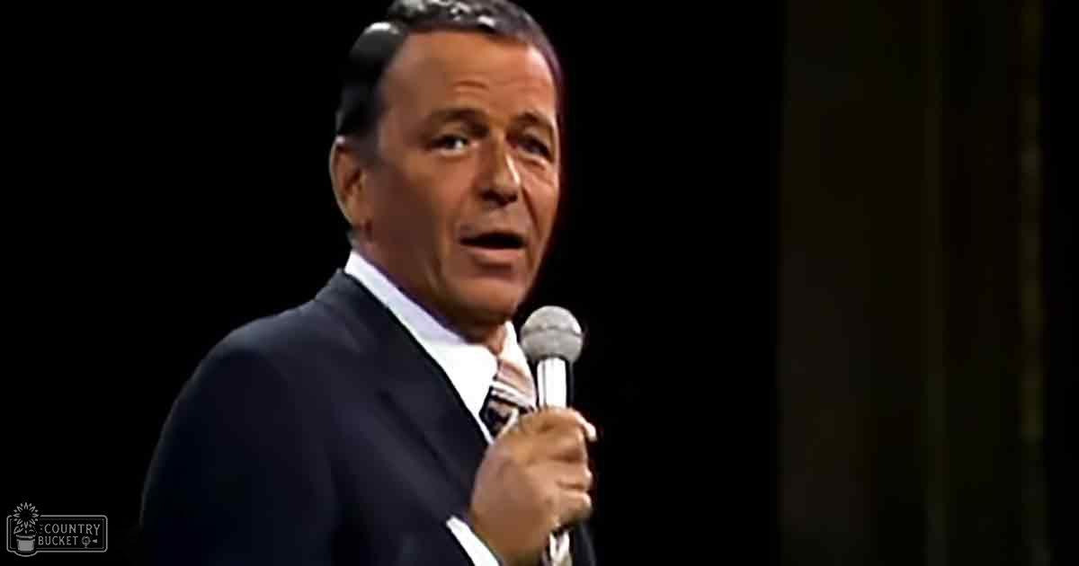 A middle-aged adult male in a dark suit jacket with a striped patterned tie, performing on stage at an indoor venue. Dark curtains and spotlight suggest a theatrical or concert setting.