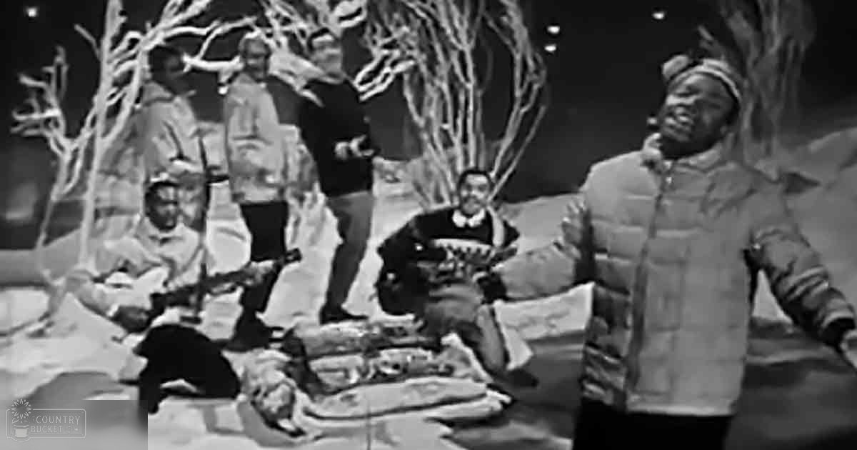 A group of individuals dressed for winter in an outdoor setting with bare trees and snow-covered ground under artificial stage lighting.
