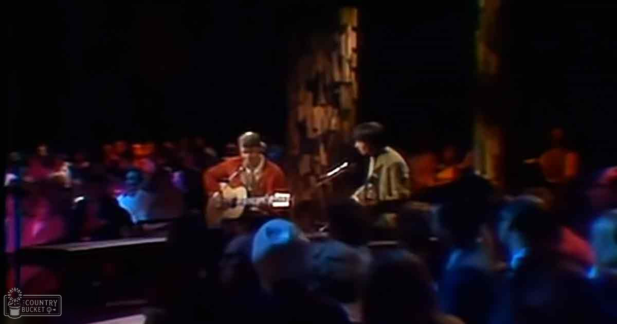 Musical performance indoors with a male performer in a red patterned jacket, audience in casual attire, piano on stage with dimly lit ornate background.