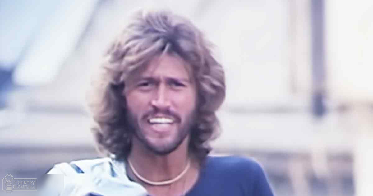 A young adult male stands outdoors, wearing a relaxed fit cotton T-shirt with a thin necklace visible around his neck. The setting appears to be an outdoor environment with blurred background structures that might suggest a bridge or similar large structure in the distance.