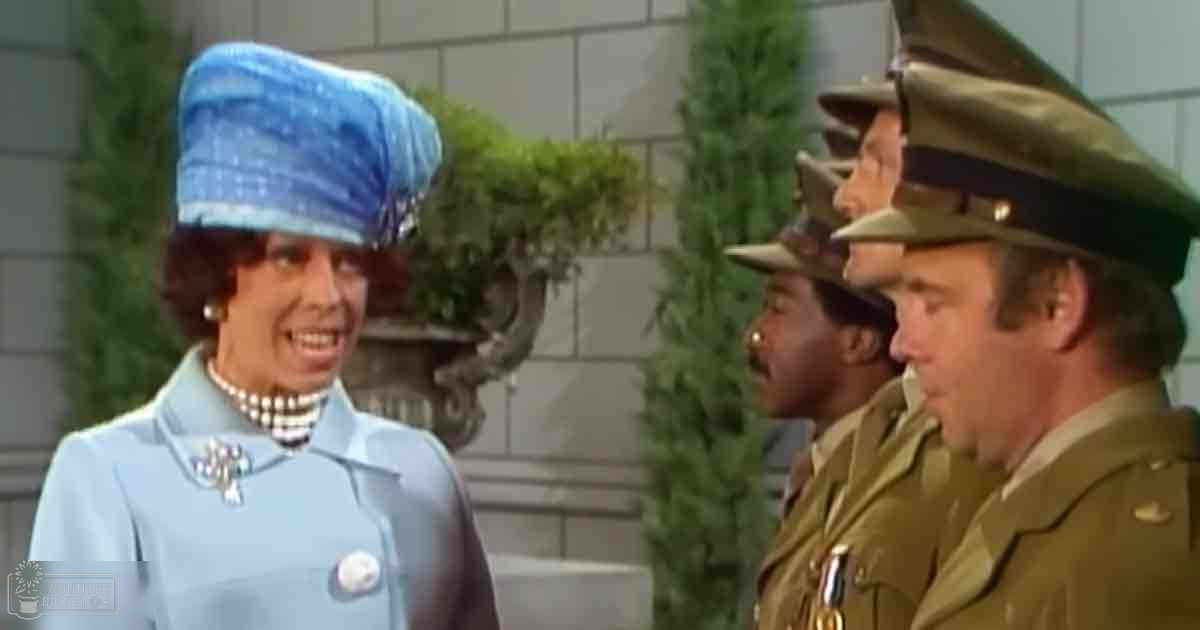 A woman in a light blue outfit with a matching hat and pearl necklace stands indoors, possibly at a formal event; men in olive green military uniforms are also present.