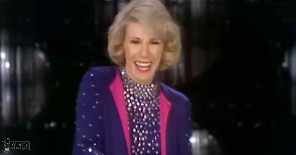 A middle-aged adult female wearing a fitted, tailored blue blazer with pink accents and small white dots or stars from the mid-20th century stands against a dark indoor background suggesting a stage or performance area.