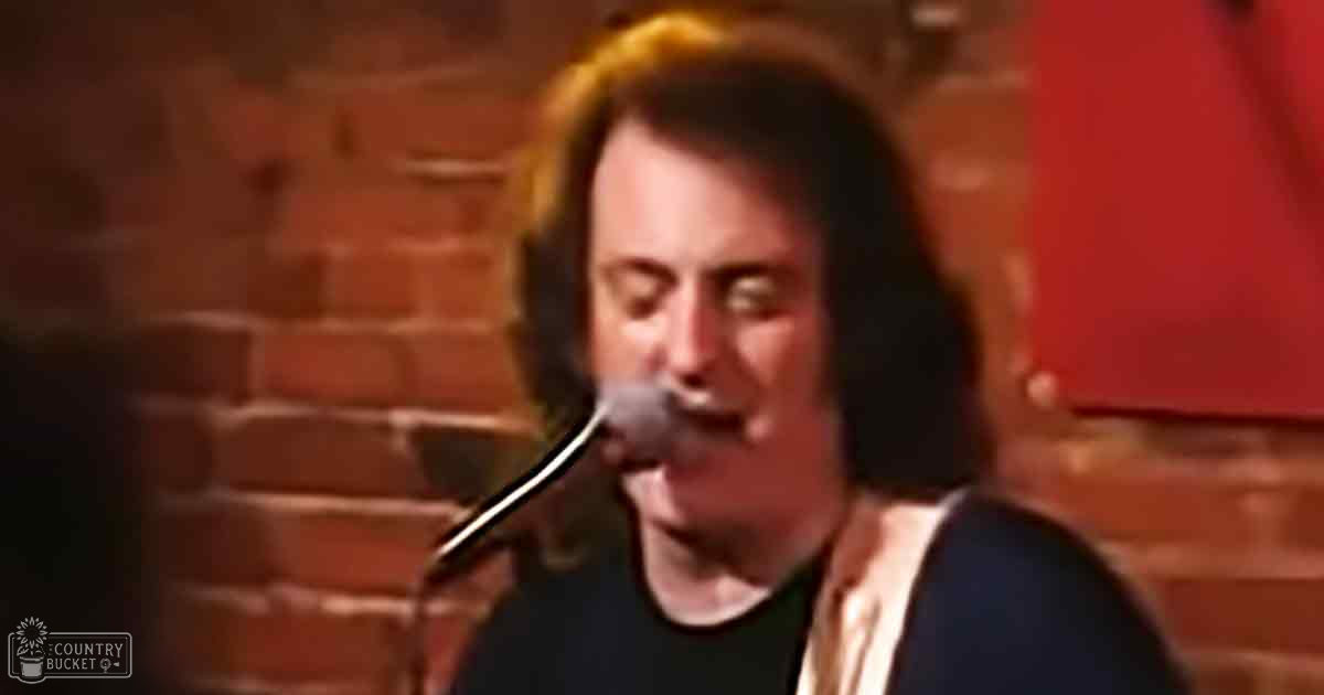 An adult male singer wearing a black t-shirt with a white strap over his shoulder, singing into a microphone in an indoor setting with brick walls under warm lighting.